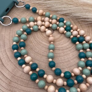 Könnte beinhalten: Eine tealfarbene Handyhülle mit einer Handy-Kette aus Holzperlen. Der Riemen hat abwechselnde Farben aus Teal, Hellgrün und Naturholzperlen. Der Riemen ist mit silbernen Metallringen an der Handyhülle befestigt.