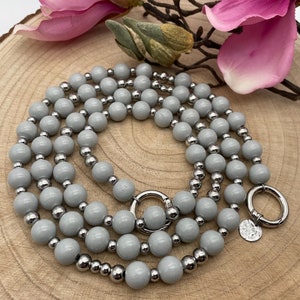 Może przedstawiać: Szary naszyjnik z koralików z srebrnymi akcentami metalowymi. Naszyjnik ma okrągłe zapięcie i srebrny zawieszkę.