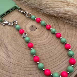 Puede incluir: Una correa de teléfono con cuentas con una funda de teléfono verde claro. La correa presenta cuentas rosas neón y verde claro alternas, conectadas por cuentas de metal plateado. La correa está sujeta a la funda del teléfono con cierres metálicos plateados.