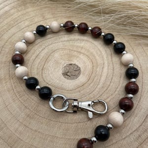 Puede incluir: Un cordón con cuentas de madera en tonos naturales, negros y marrón oscuro, intercaladas con pequeñas cuentas de metal plateado. El cordón tiene un cierre de metal plateado.