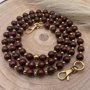 May include: A layered necklace featuring dark brown wooden beads and gold-colored metal accents. The necklace has a clasp closure and is displayed on a wooden surface. The beads are round and evenly spaced, creating a simple yet elegant design.