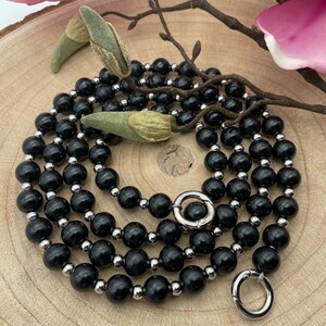 Könnte beinhalten: Eine schwarze Perlenkette mit silbernen Akzenten und zwei silbernen Verschlüssen. Die Kette liegt auf einer Holzoberfläche.