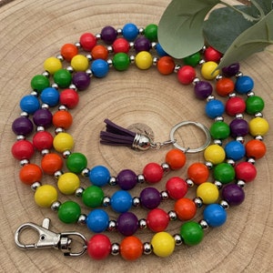 Può includere: Un laccio con perline arcobaleno con un portachiavi argentato e un fiocco viola. Il laccio è fatto di perline di legno nei colori dell'arcobaleno.