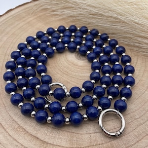 Puede incluir: Un collar de cuentas azul oscuro con detalles plateados y un cierre plateado. El collar está enrollado sobre una superficie de madera.