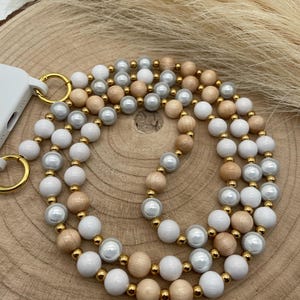 Może przedstawiać: Biały, beżowy i złoty pasek do telefonu z koralikami i złotym zapięciem. Pasek wykonany jest z drewnianych koralików, pereł i złotych koralików dystansowych.