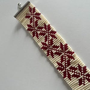 Puede incluir: Un brazalete de cuentas blancas con un patr&oacute;n geom&eacute;trico rojo. El brazalete tiene un cierre plateado.