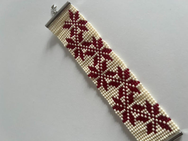 Puede incluir: Un brazalete de cuentas blancas con un patr&oacute;n geom&eacute;trico rojo. El brazalete tiene un cierre plateado.