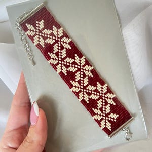 Puede incluir: Un brazalete de cuentas rojas y blancas con un patr&oacute;n de copo de nieve. El brazalete tiene un cierre plateado.