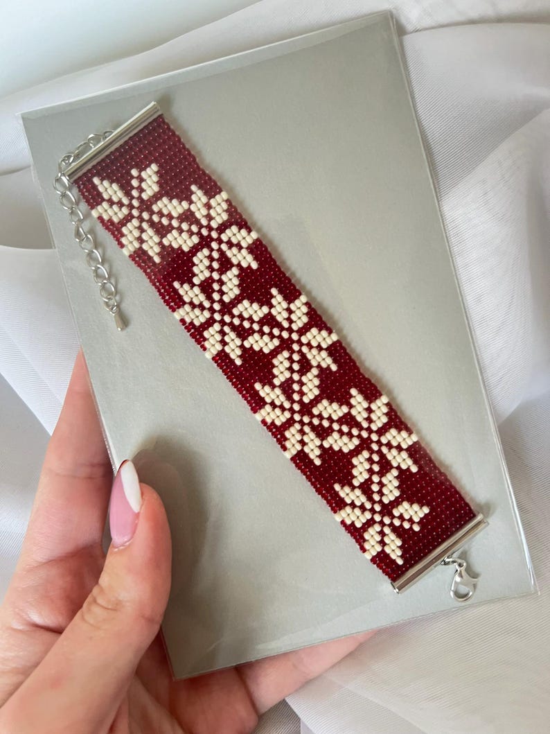 Puede incluir: Un brazalete de cuentas rojas y blancas con un patr&oacute;n de copo de nieve. El brazalete tiene un cierre plateado.