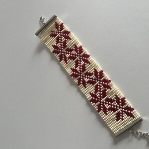 Puede incluir: Un brazalete de cuentas blancas con un patr&oacute;n floral rojo y cierres plateados.