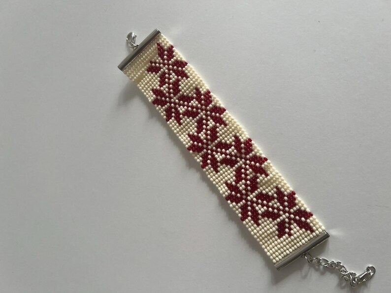 Puede incluir: Un brazalete de cuentas blancas con un patr&oacute;n floral rojo y cierres plateados.