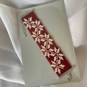 Puede incluir: Un brazalete de cuentas rojas y blancas con un patr&oacute;n de copo de nieve. El brazalete tiene un cierre plateado.