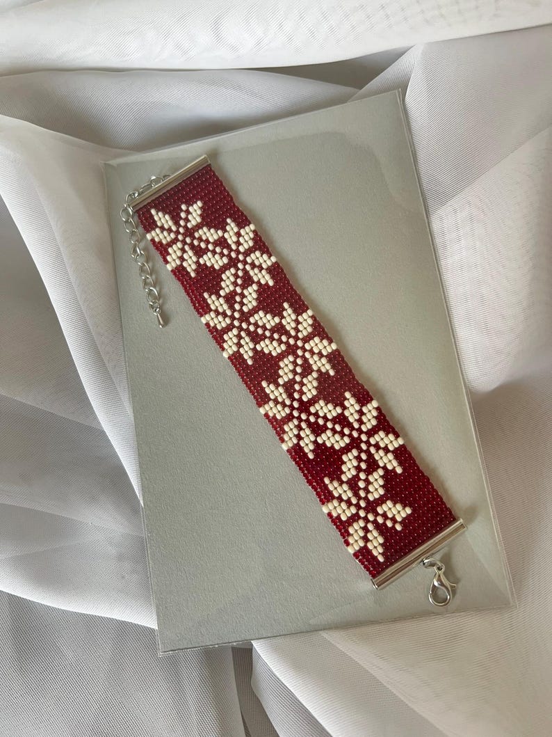 Puede incluir: Un brazalete de cuentas rojas y blancas con un patr&oacute;n de copo de nieve. El brazalete tiene un cierre plateado.