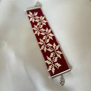 Puede incluir: Un brazalete de cuentas rojas y blancas con un patr&oacute;n de copo de nieve. El brazalete tiene un cierre plateado.