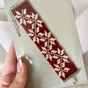 Puede incluir: Un brazalete de cuentas rojas y blancas con un patr&oacute;n de copo de nieve. El brazalete tiene un cierre plateado.