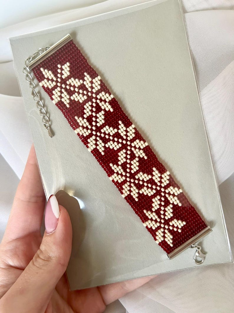Puede incluir: Un brazalete de cuentas rojas y blancas con un patr&oacute;n de copo de nieve. El brazalete tiene un cierre plateado.
