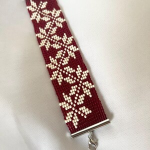 Puede incluir: Un brazalete rojo de cuentas con un patr&oacute;n de copo de nieve blanco. El brazalete tiene un cierre plateado.