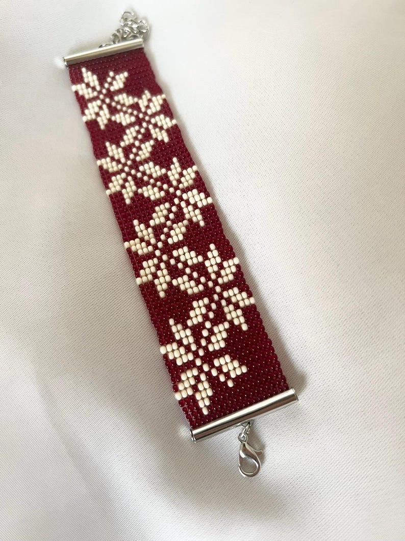 Puede incluir: Un brazalete rojo de cuentas con un patr&oacute;n de copo de nieve blanco. El brazalete tiene un cierre plateado.