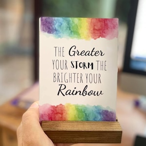 May include: A white card with a rainbow watercolour background and the text "The greater your storm the brighter your rainbow". The card is on a wooden stand.