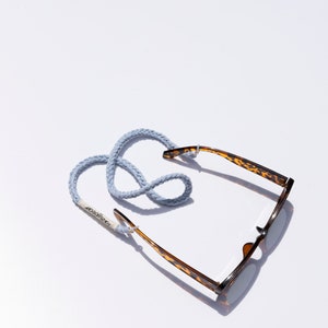 May include: Tortoise shell sunglasses with a light blue braided cord attached to the arms. The cord has a white tag with the text "#goodvibes" printed on it.