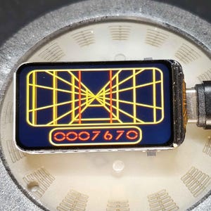 Puede incluir: Un pequeño dispositivo rectangular con una pantalla que muestra un gráfico amarillo y rojo de estilo retro, conectado a un cable negro. La pantalla muestra un diseño similar a una cuadrícula con el número "0007670".