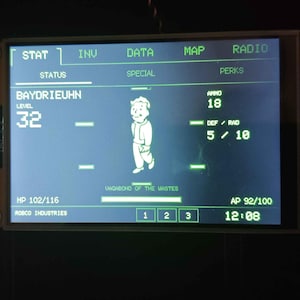 Puede incluir: Una pantalla digital rectangular que muestra una interfaz verde sobre negro con el texto STAT, INV, DATA, MAP, RADIO y ROBCO INDUSTRIES. La pantalla muestra un gráfico de personaje, nivel 32, munición 18, HP 102/116, AP 92/100 y 12:08.