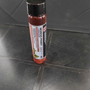 Puede incluir: Un vial cilíndrico lleno de líquido rojo, con una tapa negra de rosca y una etiqueta blanca. La etiqueta muestra el texto Umbrella Corporation, Nemesis Project, T-Virus B.O.W. y Danger: Extremely Aggressive B.O.W. con iconos de peligro amarillos.