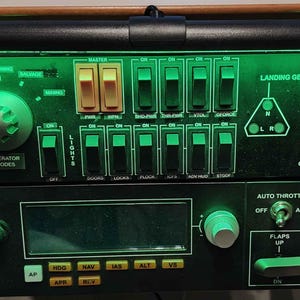 May include: A black control panel with green illuminated buttons, switches, and dials. The panel includes labels such as "Quantum," "Lights," and "Landing Gear." There are also yellow and green toggle switches and a digital display.