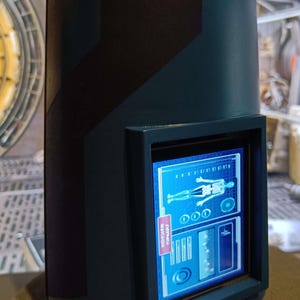May include: A dark blue, cylindrical device with a black frame around a screen displaying a blue and white graphic of a human skeleton with a medical symbol. The screen also displays text that reads "SCANNING" and "PRIORITY".
