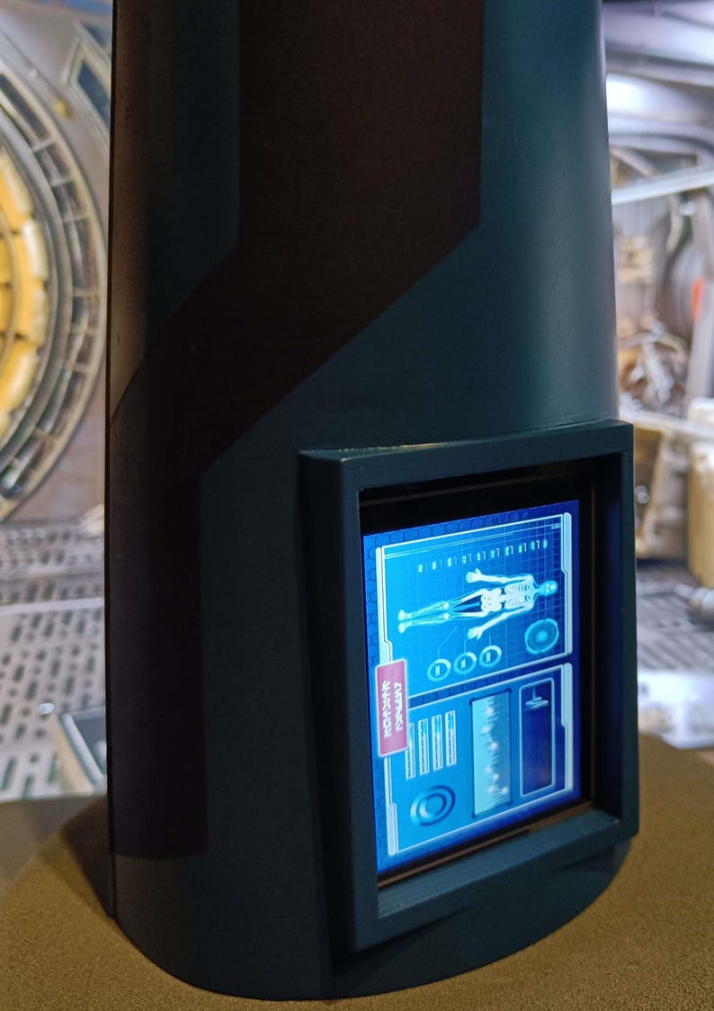 May include: A dark blue, cylindrical device with a black frame around a screen displaying a blue and white graphic of a human skeleton with a medical symbol. The screen also displays text that reads "SCANNING" and "PRIORITY".