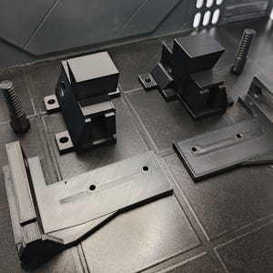 Könnte beinhalten: Eine Ansammlung schwarzer, 3D-gedruckter mechanischer Teile, darunter Halterungen, Blöcke und Schrauben. Die Komponenten sind auf einer dunklen, strukturierten Oberfläche angeordnet und zeigen ihre geometrischen Formen und ihr industrielles Design.