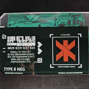 Op de afbeelding: Een rechthoekige kaart met een zwart midden en een doorzichtige plastic rand. Het zwarte gedeelte bevat witte tekst, een streepjescode en een oranje symbool in een witte vierkant. De kaart heeft ook een groen printplaatontwerp.