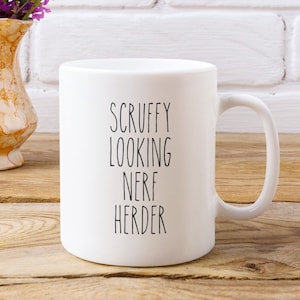 Puede incluir: Taza de cerámica blanca con asa curva. La taza presenta el texto "SCUFFY LOOKING NERF HERDER" en negro, centrado verticalmente. La taza está sobre una superficie de madera con un jarrón decorativo y flores en el fondo.
