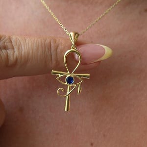 May include: A gold-coloured pendant necklace featuring an Ankh symbol with a blue gemstone eye. The necklace is worn around the neck. The Ankh pendant is held by a hand.
