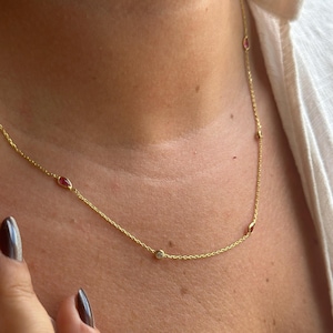 May include: A delicate gold necklace featuring small, oval-shaped red gemstones and tiny round clear stones. The necklace is worn against a white shirt, showcasing its fine chain and minimalist design. The jewelry is a subtle yet elegant accessory.