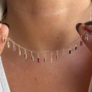 May include: A gold-coloured necklace with small, rectangular gemstones in various colours. The gemstones are suspended from a fine chain, creating an elegant design. The necklace is being held up, showcasing its details.