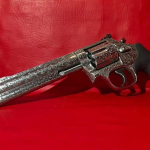 May include: A silver revolver with intricate floral engravings. The gun features a long barrel and a black grip. The firearm is set against a red background, highlighting the details of the metalwork.