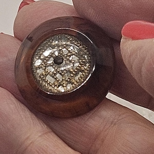 Op de afbeelding: Een ronde, vintage knoop met een donkerbruine, glanzende buitenring. Het midden heeft een zilver- en goudkleurig filigraanontwerp. De knoop wordt in een hand met rode nagellak gehouden.