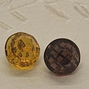 May include: Two faceted, round buttons. One is amber-colored, the other is dark brown. Both buttons have a geometric pattern of small, angled facets across their surfaces. The buttons are set against a light-colored background.