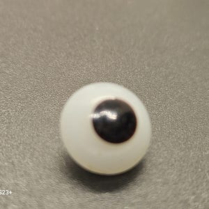May include: A single, round, white eye with a dark brown iris. The eye is set against a gray background. The eye is a craft supply, likely for dolls or other art projects.