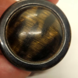 May include: A round, dark brown button with a metallic rim. The button's center features a swirling pattern of dark brown and gold tones. The button is likely made of wood or a similar material.