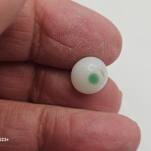 Puede incluir: Una pequeña cuenta redonda blanca con un punto verde. La cuenta se sostiene entre los dedos, mostrando su tamaño y detalle. La superficie de la cuenta parece lisa, con un ligero brillo.