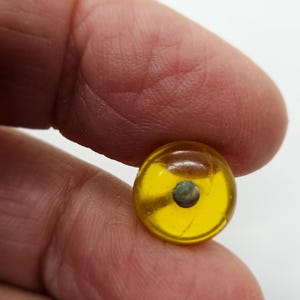 May include: A small, round, transparent yellow bead with a dark, circular inclusion in the center. The bead is held between two fingers against a white background. The bead is approximately 0.5 inches in diameter.