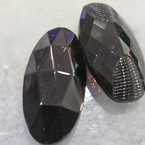 Puede incluir: Dos gemas oscuras, facetadas, de forma ovalada. Las gemas tienen un diseño geométrico con múltiples facetas que capturan la luz, creando un juego de colores. Una gema tiene un extremo texturizado.