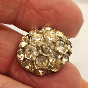 May include: A round, silver-toned button covered in numerous clear, sparkling rhinestones. The rhinestones are closely set together, creating a dense, textured surface. The button is held between fingers against a neutral background.