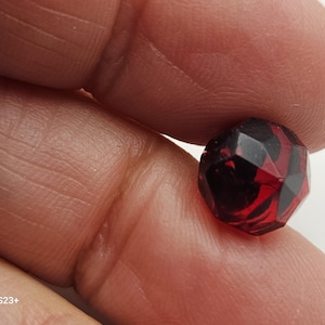 May include: A faceted, round bead with a deep red and black color scheme. The bead's surface is multi-faceted, creating a play of light and shadow. The bead is held between fingers, showcasing its size and detail.