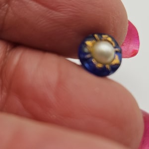Puede incluir: Un pequeño botón redondo con una perla central rodeada por un borde azul oscuro con detalles dorados. El botón se sostiene entre los dedos, mostrando su intrincado diseño. El botón es un hallazgo de joyería.