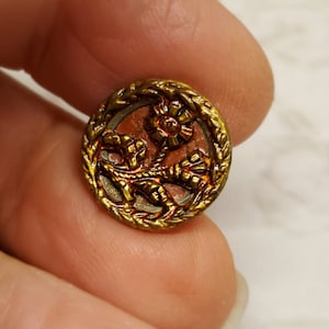 May include: A round, antique button with a floral design. The button features a gold-colored metal frame with a detailed floral pattern in the center. The center of the button has a reddish-brown hue. The button is held between two fingers.