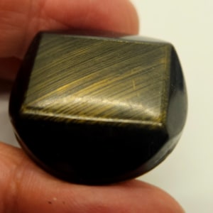 May include: A dark, faceted stone with a rectangular top and rounded base. The top surface has a golden, striated pattern. The stone is held in a hand, suggesting its size and shape.