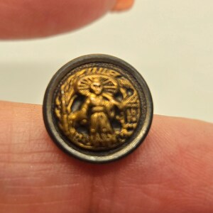 May include: A round, antique button with a detailed gold-colored relief design. The central figure is a person with outstretched arms, surrounded by wheat and a building. The button has a dark, metallic border.